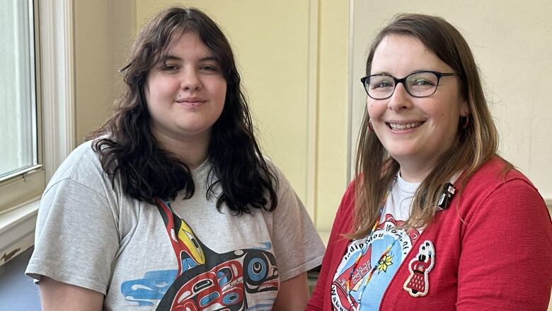 A young person with dark, curly hair stands next to an older woman wearing glasses and a red cardigan. 