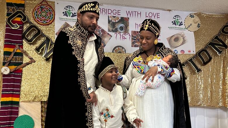 A family stands in front of decorative gold cloth hanging on the wall. They are wearing traditional black and gold cloaks. 