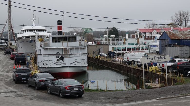 Two large boats floating in a dock area, surrounded by parked cars and workshops.