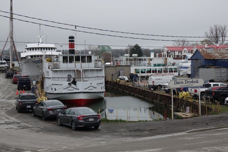 Two large boats floating in a dock area, surrounded by parked cars and workshops.