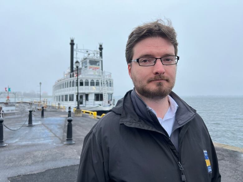 A man with brown hair and glasses. Behind him is a white, paddle-steamer style boat. It's a very foggy, grey day.