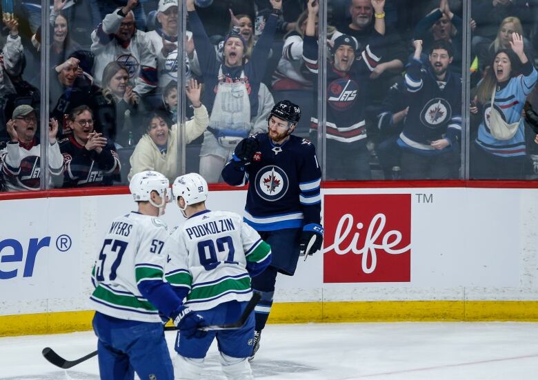 Two Canucks players watch while a Winnipeg player pumps his fist in front of many cheering Jets fans behind the glass.