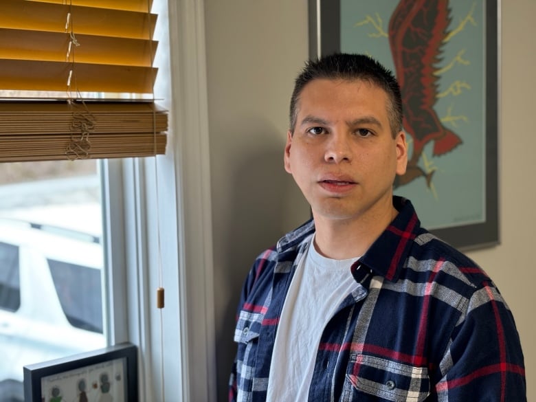 A man wearing a plaid shirt stands beside a window.