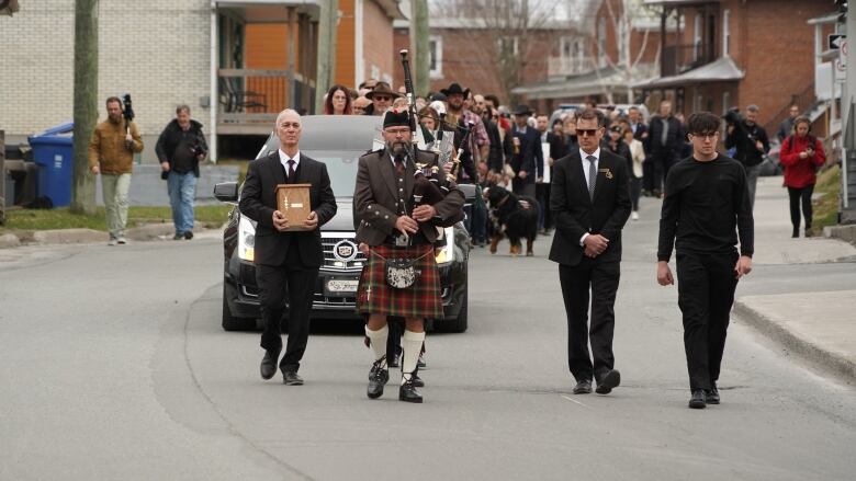 A funeral procession lead by a bagpipe player and Jacob Flickinger's father holding the urn with his ashes. 