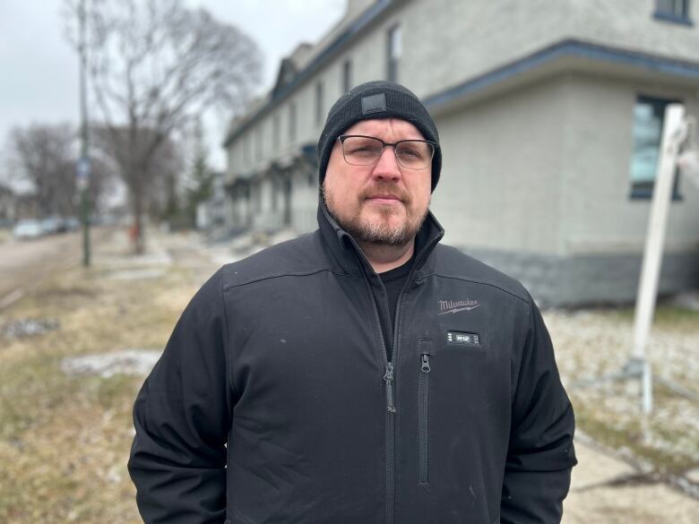 A man wearing a black had and jacket is standing outside. A row of brick townhouses can be seen behind him.