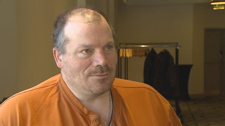 A balding man wearing an orange shirt looks at the camera.