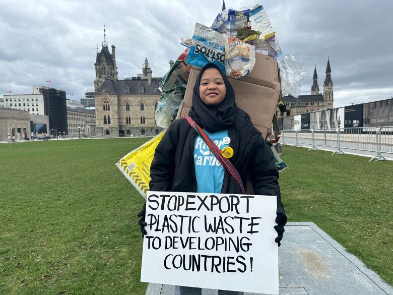 A teen girl dressed in a costume made of plastic waste stands on a lawn outdoors and holds a protest sign.