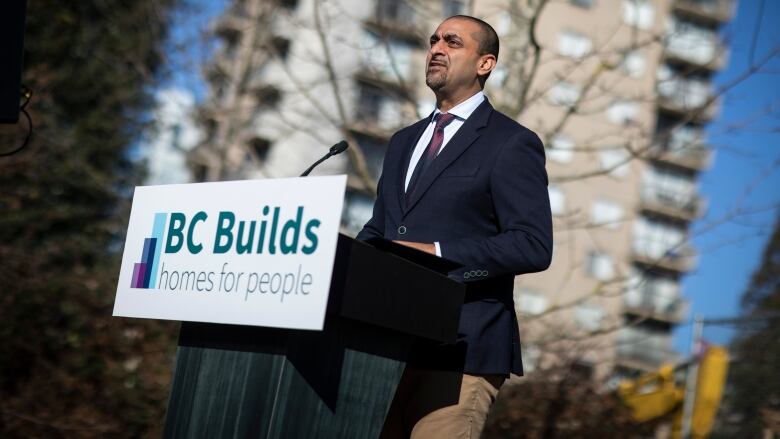 A South Asian man stands at a podium marked 'B.C. Builds', with an apartment building behind him.