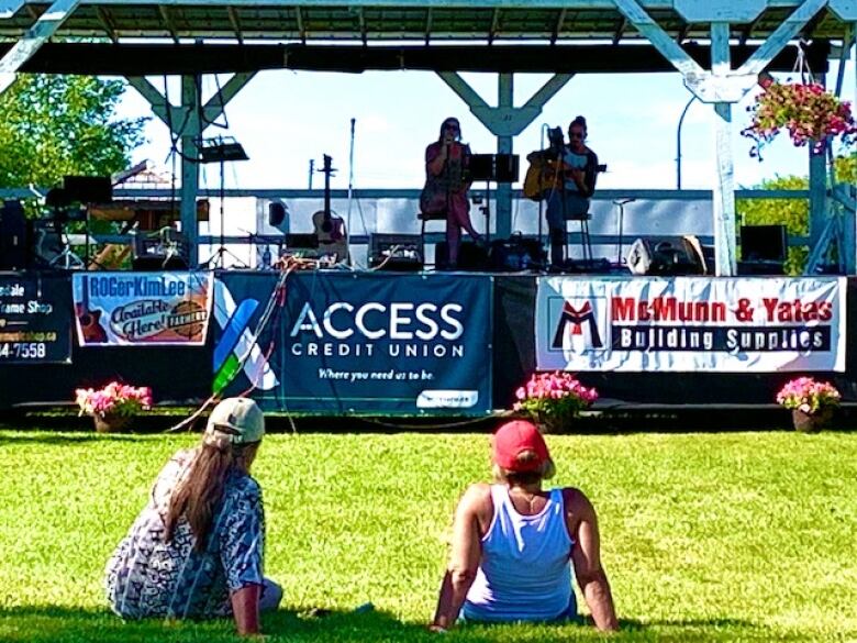 Two people lay on the grass in front of a stage while musicians play.