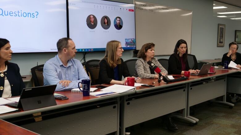 People sit at a long table with a PowerPoint presentation on the screen in the background 