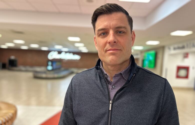 Man in a dark grey zip-up sweatshirt stands in an airport arrivals lounge.