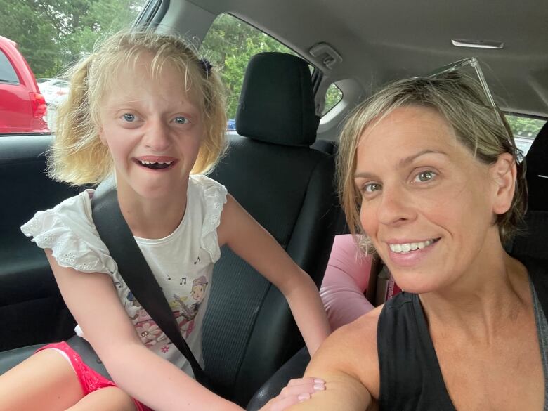 A young girl with blonde hair is in the car with her mother. They are both smiling.