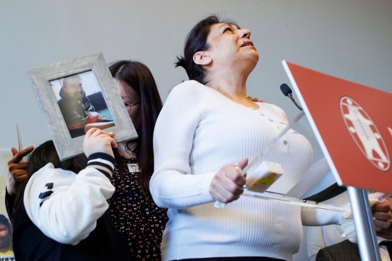 A woman wearing a white shirt is seen speaking at a podium. Beside her are two other people, shielding their faces with a framed photograph of a man.