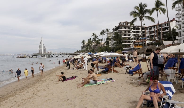 Sunbathers lie on a tropical beach. There are palm trees and a hotel in the background. 