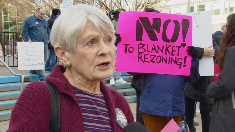 a woman speaking in front of a crowd of people. a person holds a bright sign that says 