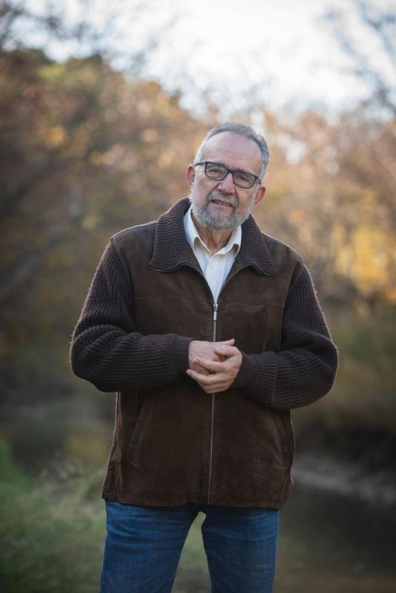 A man stands and looks at the camera in front of a soft-focus background of autumn trees.