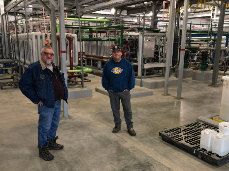 Two men stand in front of large and complex equipment and pipes.