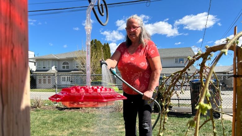 A woman wearing a red shirt and black pants waters her garden with a hose