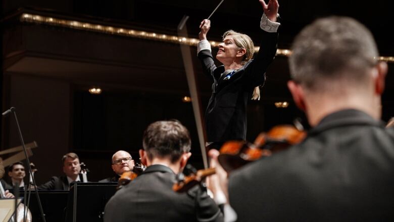 A conductor joyfully lifts her arms in front of an orchestra.