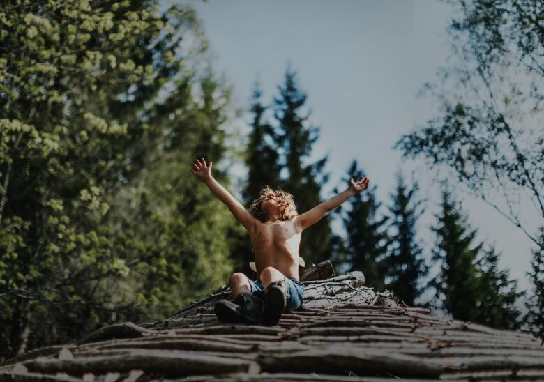 A child sitting on a roof thatched with twigs in the woods raises their arms joyfully.