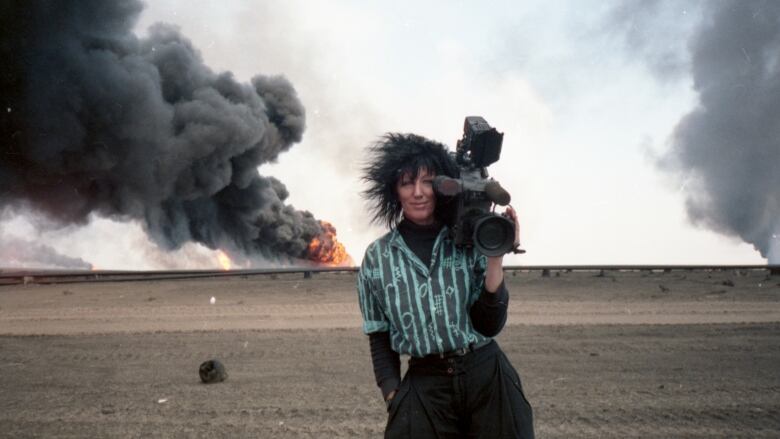 A smiling woman with wild black hair stands in front of what looks like an explosion belching smoke in the background. 