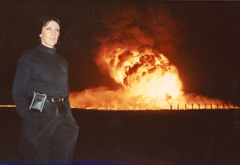 A slightly smiling woman stands in front of a large fire at night. 