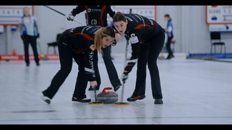 Two teenage girls brush furiously in front of a curling rock on a rink. 