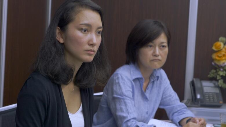 A woman with a vacant expression, left, sits next to a frowning woman. Both are sitting at a table in an office. 