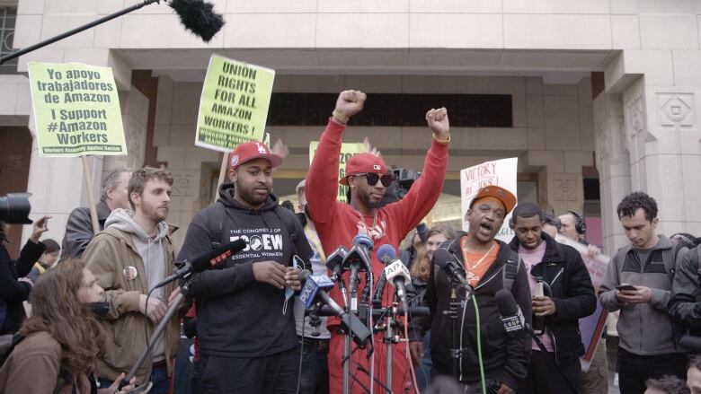 A man standing in front of a stand full of microphones holds both his fists in the air. Behind him a crowd of people cheer. Some hold signs that say 'Union rights for all Amazon workers.'