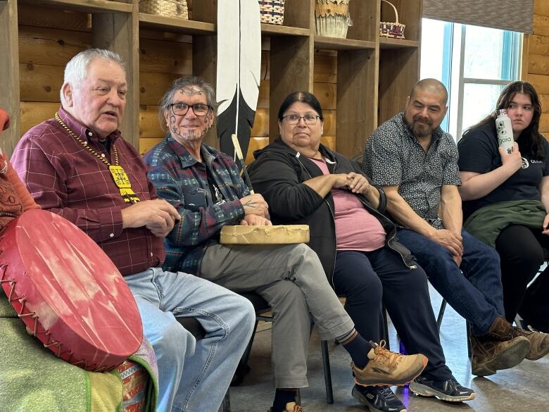 A group of people sit on folding chairs in a room with wooden walls.