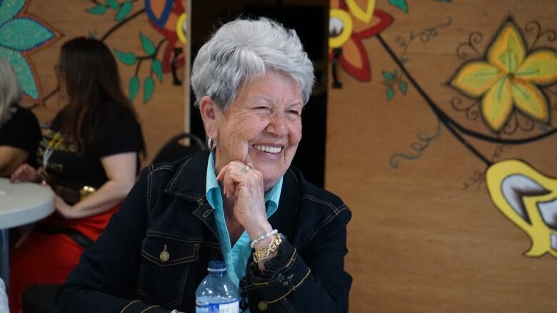 A woman with short, white hair sits at a table smiling.