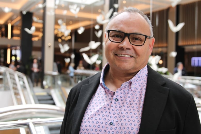 A man with short grey hair, black framed glasses, black jacket and pink shirt with blue dots smiles while standing in a hotel lobby. 