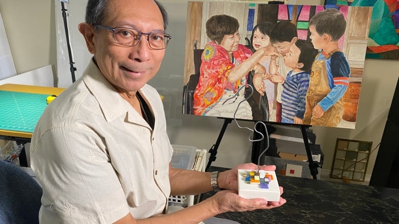 A man in glasses holds a wire sculpture in his hand, the curves representing a smaller figure bending over the hand of a taller figure. A painting behind him shows young children kissing the hand of a senior citizen.