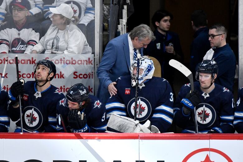 A man in a suit talks to a goaltender seated on the team's bench.