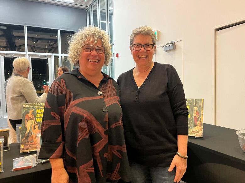 Two women are grinning and standing in front a table featuring pulp fiction novels.