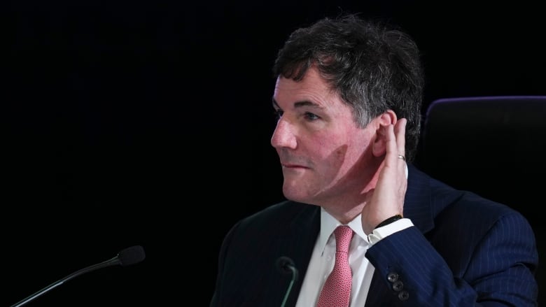 A white man wearing a red tie holds his hand to his ear while sitting in front of mics.