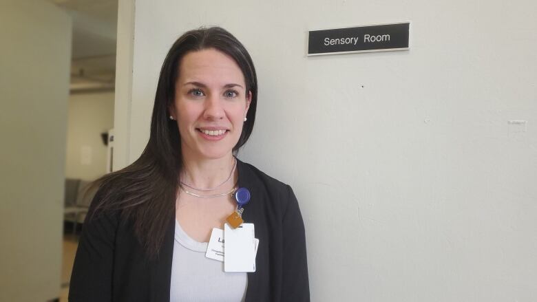 A woman with black hair wearing a white shirt and a black blazer with ID tags pinned to her lapel smiles at the camera while standing in front of a door with a tag that reads 