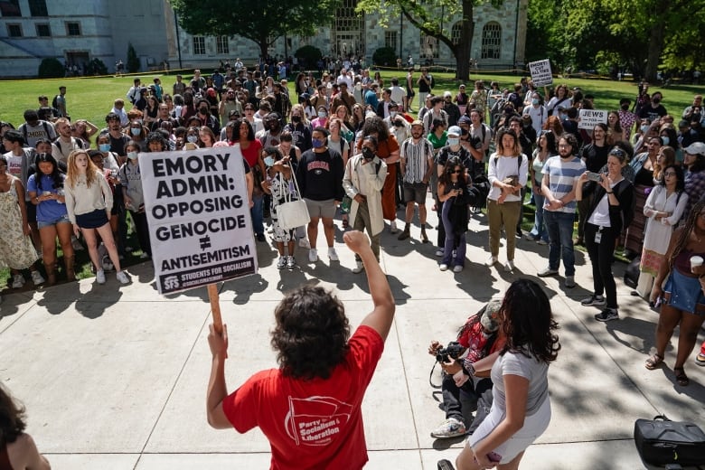 A person in a red t-shirt faces a crowd on a university campus while holding up a protest sign reading 