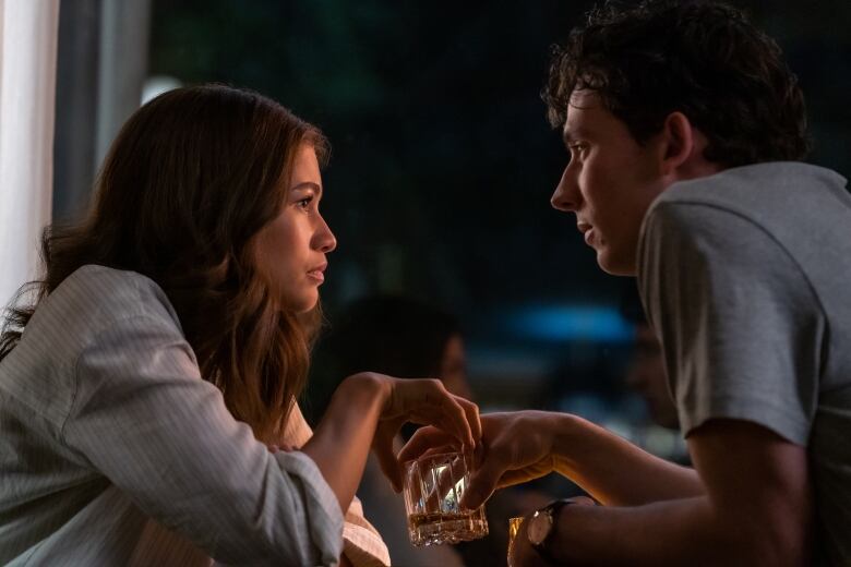 A woman and man sit across from one another. The man is holding a glass of whisky and the woman lightly puts her hands on the same glass while the two look into one another's eyes. 