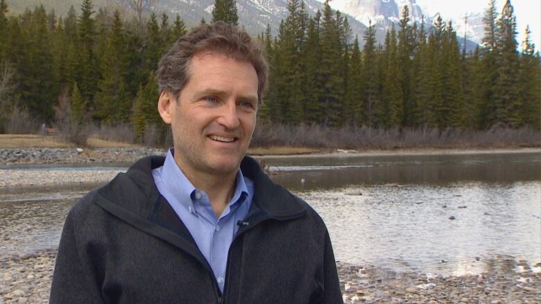 A man wearing a jacket stands in front of a river.