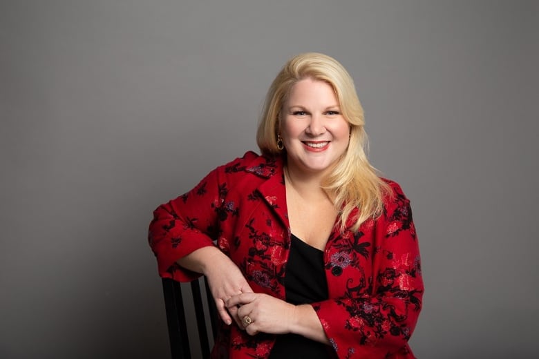 Smiling blonde woman, wearing a red blazer with a floral pattern, poses in a chair in front of a grey studio backdrop.