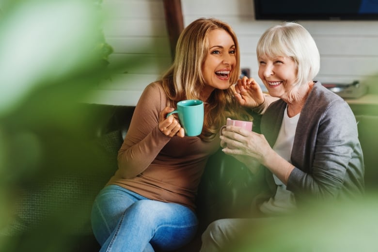 Two women sitting on a sofa, holding mugs, laugh while talking to each other 