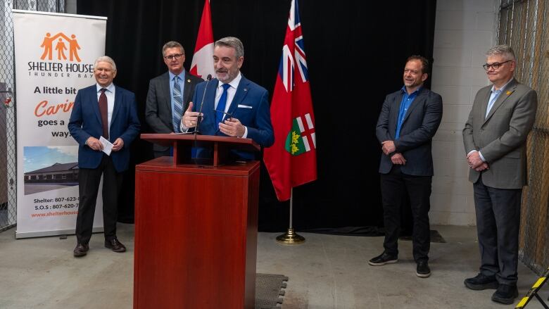 A man speaks at a podium while others stand behind and listen.