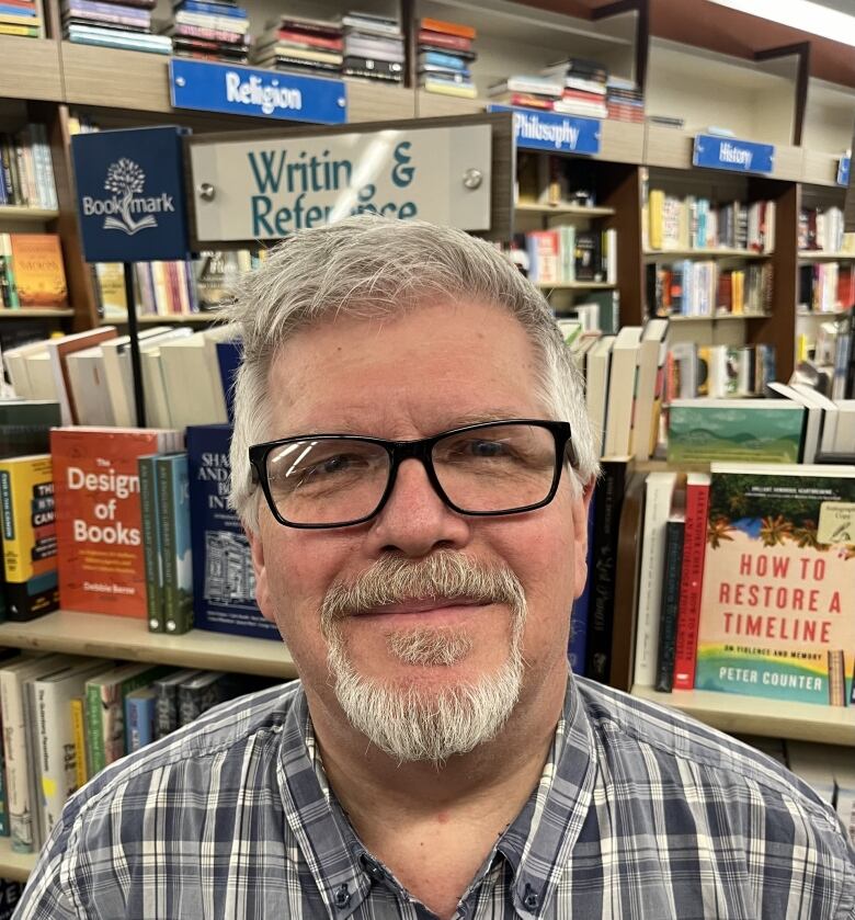 A man with grey hair and a bear stands in front of shelves of books