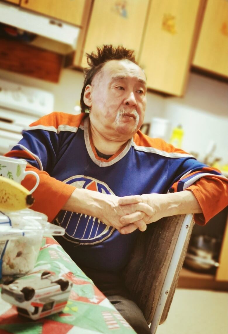 A man with a white mustache and receding black hair wearing an Edmonton Oilers jersey sitting at a dining table