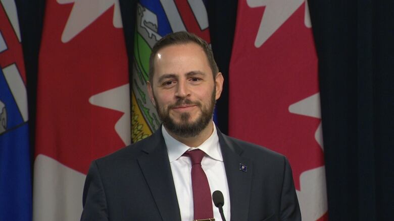 A man smiles at a microphone with Canadian and provincial flags behind him.