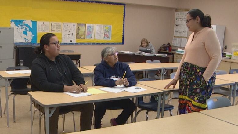 a young man and women sit at a long table while a teacher stand in front of them to answer their questions.