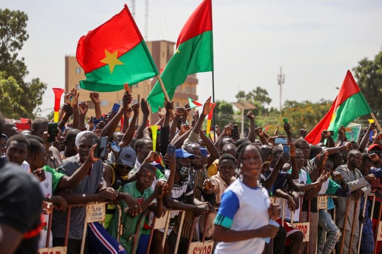 Dozens of people stand outside waving Burkino Faso flags, bright red and green with a single yellow star.