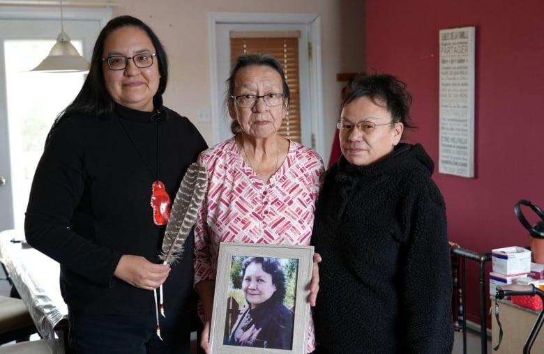 Three women look at the camera holding a photo of another woman 