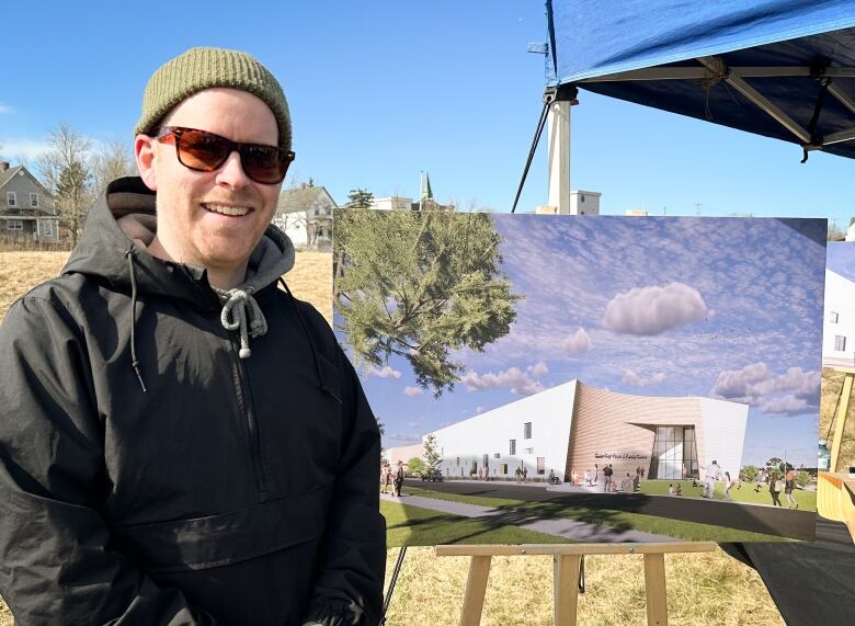 A man in a dark winter parka with a green toque smiles next to an artist's rendering of what a new building could look like.